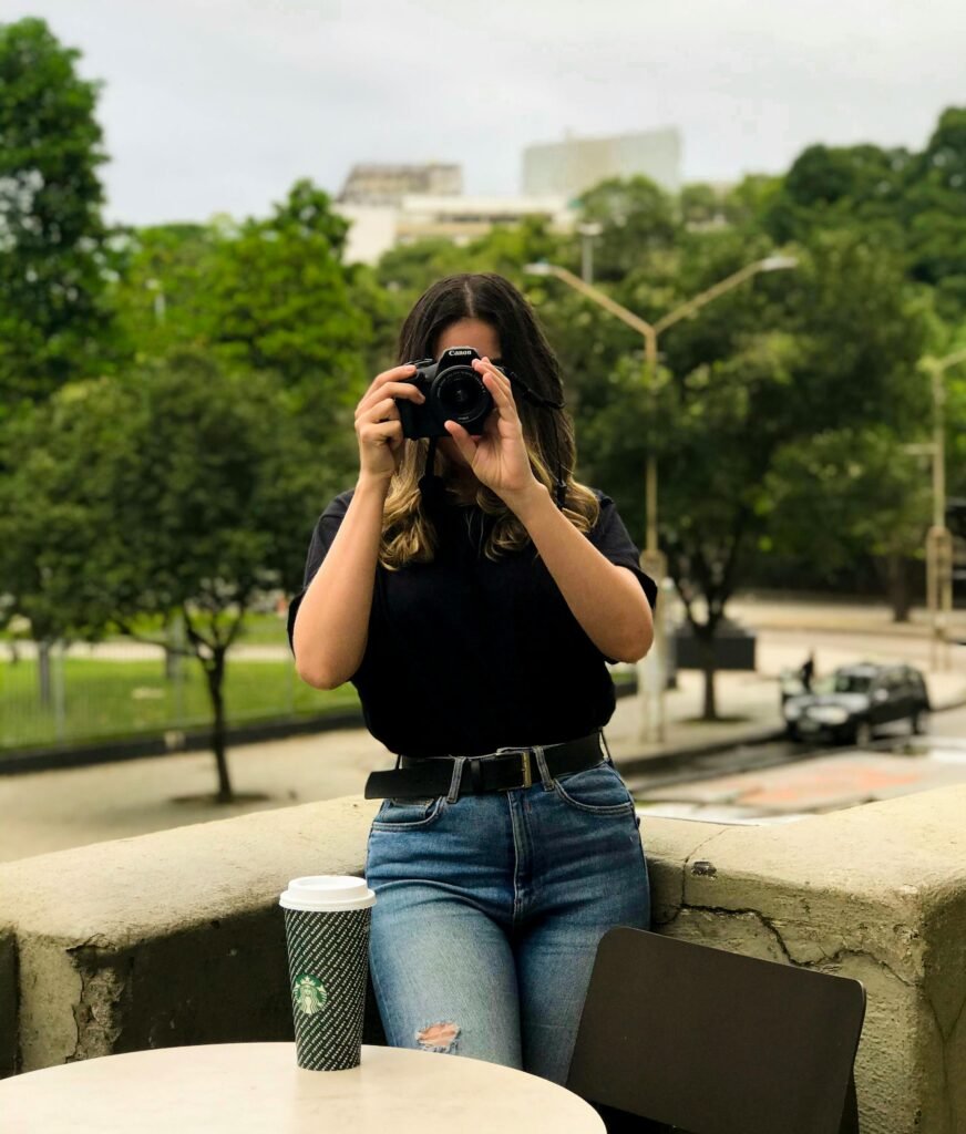 Woman in black shirt taking a photo outdoors with DSLR camera, coffee cup on table.